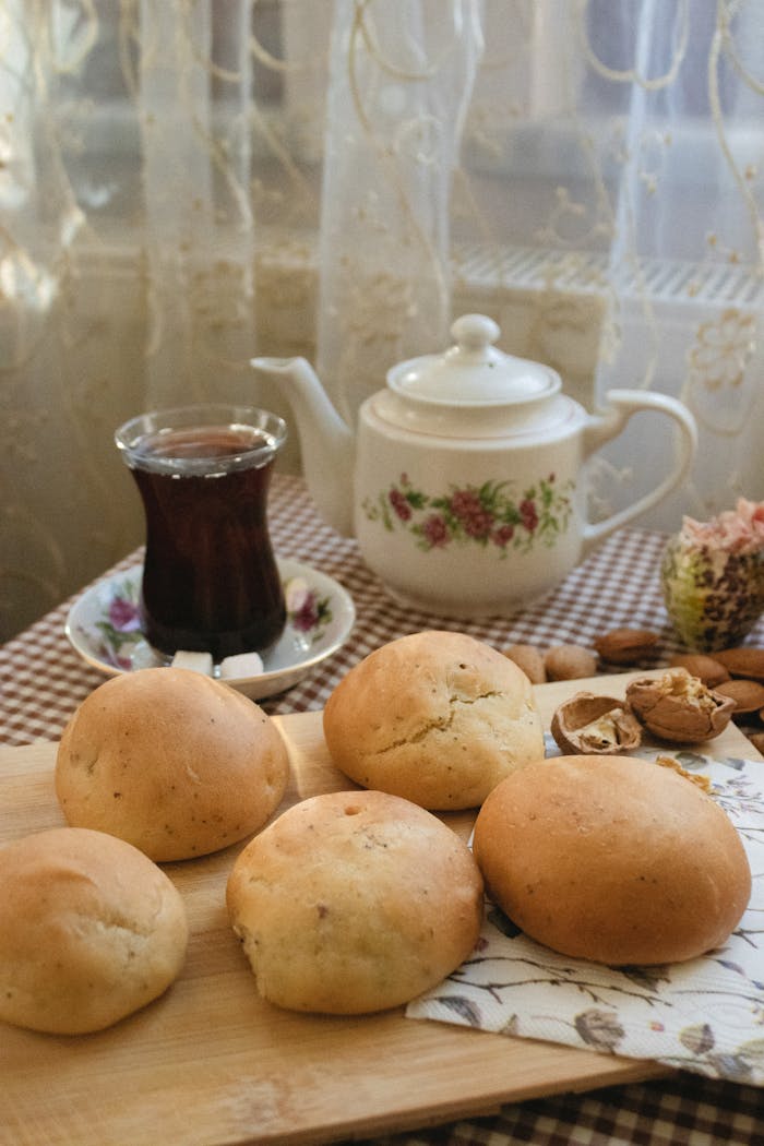 A warm morning scene featuring fresh baked rolls with a floral teapot and a glass of tea on a checkered tablecloth.